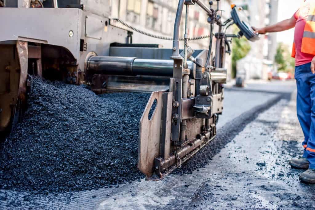 Asphalt paving machine laying fresh pavement on city road, with worker in safety gear.