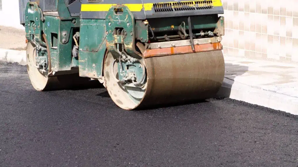Teal and yellow road roller with large metal drums compacting black asphalt pavement during construction