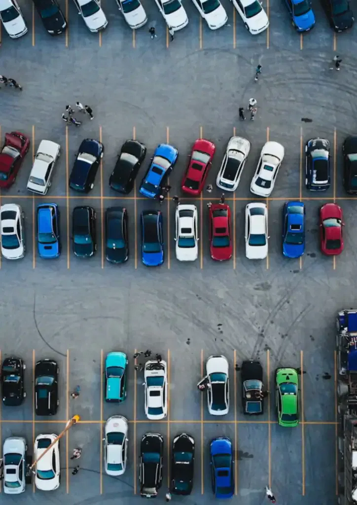 Aerial view of a busy parking lot with numerous cars in various colors including white, blue, red, black, and green, parked in marked spaces, with a few people walking around