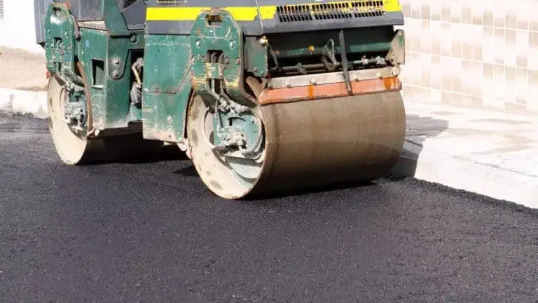 Teal and yellow road roller with large metal drums compacting black asphalt pavement during construction